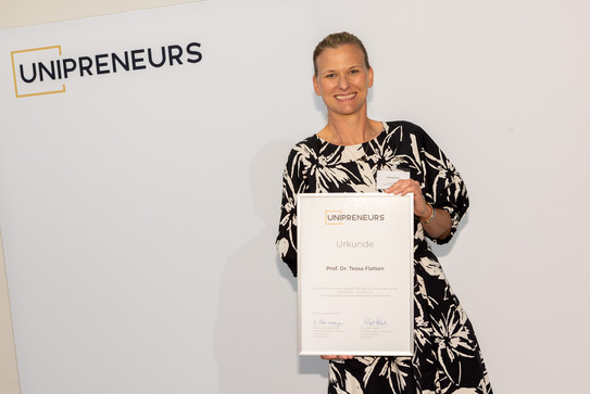Eine Frau, Prof. Tessa Flatten, hält ihre Auszeichnung mit der Aufschrift "Unipreneurs" in den Händen und zeigt sie in die Kamera. 