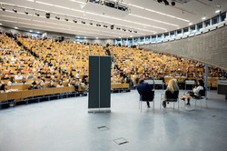 A full lecture hall. Three people are sitting on chairs in conversation.