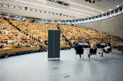 Ein voller Hörsaal. Drei Personen sitzen auf Stühlen im Gespräch.