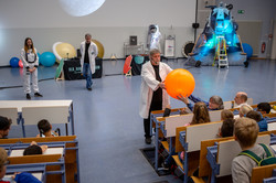 Ein Mann steht in einem vollbesetzten Hörsaal und gibt einen Wasserball, welcher einen Planeten symbolisieren soll, an im Publikum sitzende Kinder weiter.
