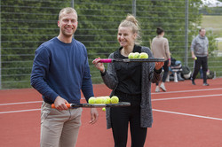 Zwei Personen halten Tennisschläger in der Hand und lächeln in die Kamera.