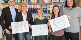 Fünf Personen stehen nebeneinander und lächeln in die Kamera. Die drei in der Mitte halten weiße Tafeln mit mathematischen Formeln in den Händen.