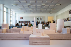 Das Bild zeigt ein Architekturmodell aus Holz mit mehreren Gebäuden. Im Hintergrund ist verschwommen der Rest des Raumes zu sehen.