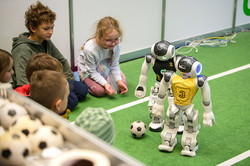 Das Bild zeigt zwei Roboter in der Ecke eines Fußballfeldes, die ihren Blick auf den Ball richten. Hinter der Linie sitzen Kinder auf dem Boden, die sich das Spiel anschauen.