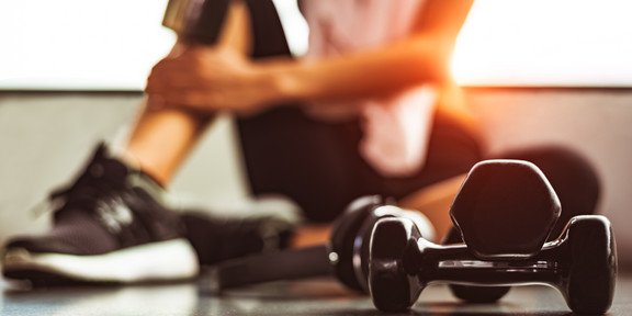 Trainingseinheit Im Vordergrund sind zwei übereinander gelegte Hanteln zu sehen. Verschwommen im Hintergrund ist unscharf eine Frau in Sportkleidung zu sehen, die mit aufgestelltem Bein auf dem Boden sitzt.