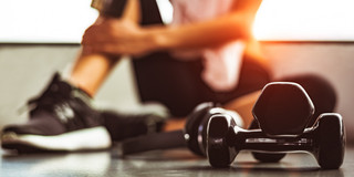 Two stacked dumbbells can be seen in the foreground. A woman in sportswear can be seen blurred in the background, sitting on the floor with her leg up.