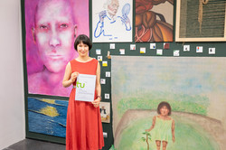 Im Zentrum des Bildes ist Dulana Tillmann zu sehen, die mit ihrem Preis-Zertifikat vor einer schwarzen Wand mit Kunstwerken steht.