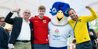 Gruppenfoto: Drei Personen posieren gemeinsam mit dem Maskottchen der Rhine-Ruhr FISU Games.