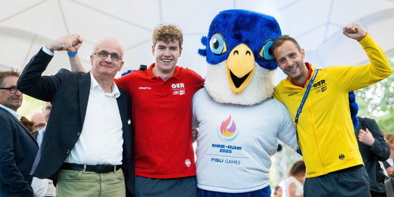 Gruppenfoto: Drei Personen posieren gemeinsam mit dem Maskottchen der Rhine-Ruhr FISU Games.