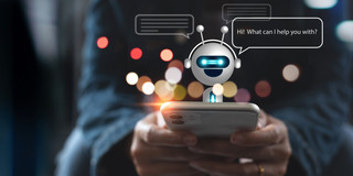 Foto: Eine Person hält ein Smartphone in der Hand, aus dem ein kleiner, lächelnder Chatbot mit Sprechblase erscheint.