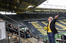 Ein Mann steht vor einer mit Erstsemesterstudierenden gefüllten Tribüne in einem Fußballstadion, hält in der rechten Hand einen Fußball und gestikuliert mit der linken Hand.