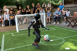 Ein humanoider Roboter kickt einen Mini-Fußball. Im Hintergrund ein kleines Tor und Zuschauer*innen.