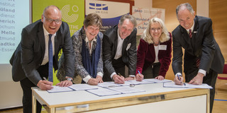 Five people lean next to each other on a table and sign a document.