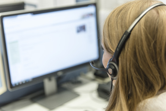 Mitarbeiterin tefefoniert mit Headset und bearbeitet eine Anfrage am Computer.