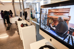 Das Foto zeigt weiße, nebeneinander Aufgestellte Podeste mit Apple Bildschirmen und schwarzen Kopfhörern. Auf dem Bildschirm selbst ist ein Mann zu sehen, der auf dem Boden zum Klavier gerichtet auf den Boden sitzt.