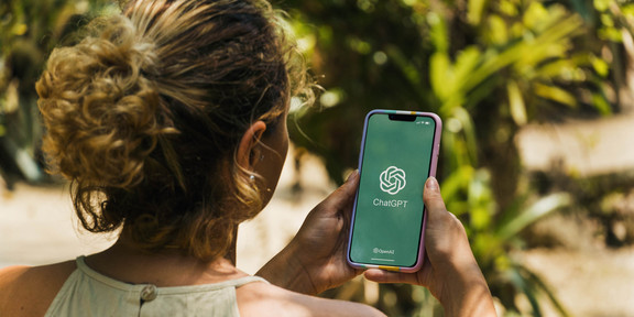 Diego_AdobeStock_580895926_Editorial_Use_Only Das Bild zeigt eine Frau von hinten mit einem Smartphone in der Hand auf dessen Bildschirm das Logo von ChatGPT zu sehen ist.