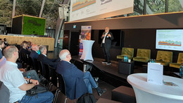 Ein Professor steht auf der Bühne und hält eine Keynote. Die Präsentation ist auf einer großen Leinwand zu sehen. Vor der Bühne sitzt Publikum.