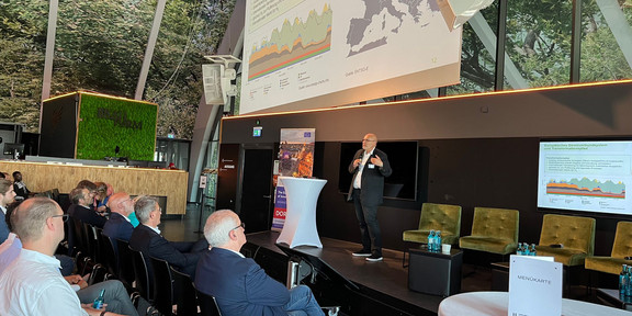 A professor stands on stage and gives a keynote speech. The presentation can be seen on a large screen. An audience sits in front of the stage.