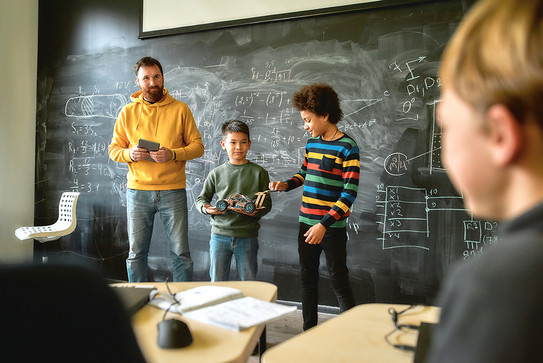 Ein Mann steht gemeinsam mit zwei Jungen vor einer Tafel in einem Klassenraum.