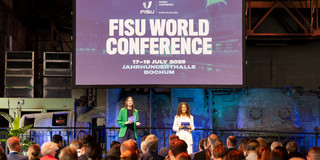 Foto: Blick auf die Bühne der FISU World Conference in der Jahrhunderthalle – Besucher*innen verfolgen sitzend das Programm.
