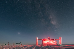 IceCube-Laboratory A red illuminated steel structure in the snow under a night sky with a lot of stars.