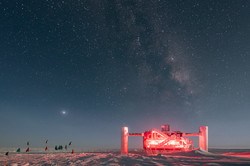A red illuminated steel structure in the snow under a night sky with a lot of stars.