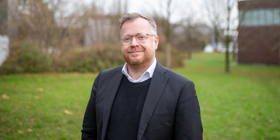 Ein Mann mit Sakko auf dem Campus vor dem Erich-Brost-Gebäude.