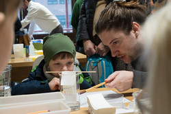 Das Bild zeigt eine erwachsene Person, die sich zu einem Kind runterbeugt, welches sich ein Experiment in einem Glas anschaut.