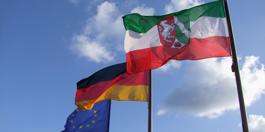 Es ist eine NRW-Flagge, eine Deutschlandflagge und eine EU-Flagge zu sehen.