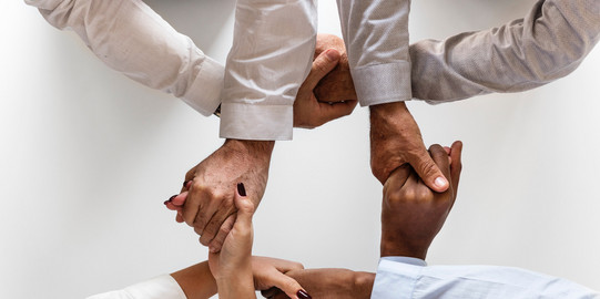 Arme von vier Menschen unterschiedlicher Hautfarbe in Business-Kleidung von oben gesehen, die Personen halten sich gegenseitig an den Händen