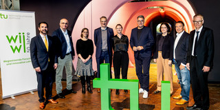 Gruppenbild von drei Frauen und sechs Männern in formeller Kleidung, die hinter einem großen, grünen TU-Logo stehen.