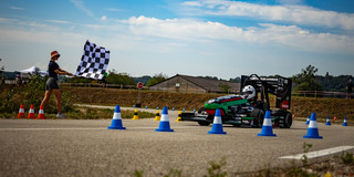 GET racing bei Formula Student France Ein kleiner Rennwagen fährt an einem sonnigen Tag über eine Rennstrecke, auf der mit bunten Sicherheitskegeln Hindernisse aufgestellt sind. Links im Bild schwenkt eine junge Frau ein schwarz-weiß-karierte Fahne.