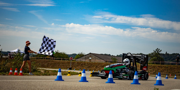 GET racing bei Formula Student France Ein kleiner Rennwagen fährt an einem sonnigen Tag über eine Rennstrecke, auf der mit bunten Sicherheitskegeln Hindernisse aufgestellt sind. Links im Bild schwenkt eine junge Frau ein schwarz-weiß-karierte Fahne.