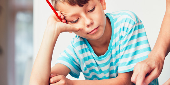 Ein Junge sitzt an einem Schreibtisch. Er schaut in ein Buch, stützt seinen Kopf auf den Arm und hält einen Stift in der rechten Hand.