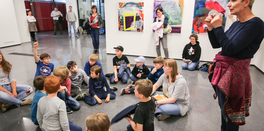 Kinder sitzen in einem Kreis auf dem Boden. An der Wand hinter ihnen hängen Bilder. Eine Frau dolmetscht für Gehörlose.