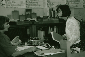 Eine Frau sitzt an einem Schreibtisch, eine andere Frau spricht mit ihr. Im Hintergrund an einer Tafel Informationen zum Standort von Büchern.
