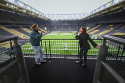 Zwei Sänger*innen, die auf der Tribüne des BVB-Stadions singen. Im Hintergrund das Fußballfeld und die leeren Tribünen des Stadions.