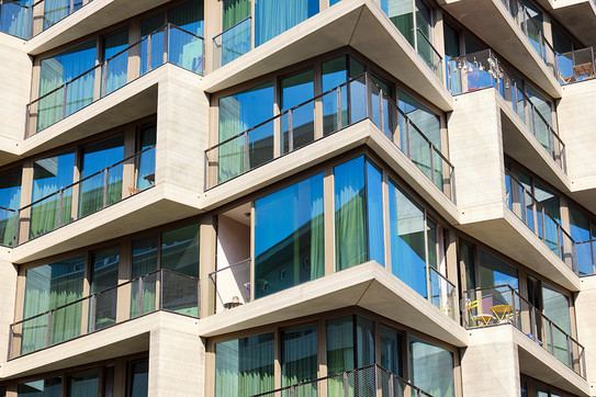 Der Bildausschnitt zeigt vier Stockwerke eines modernen Hauses mit verglasten Fassaden und architektonisch interessant gestalteten Elementen, die die Wohnungen und Stockwerke äußerlich abgrenzen.