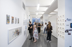 Mehrere Personen schauen sich in einer Kunstausstellung Bilder an, die an einer weißen Wand hängen.