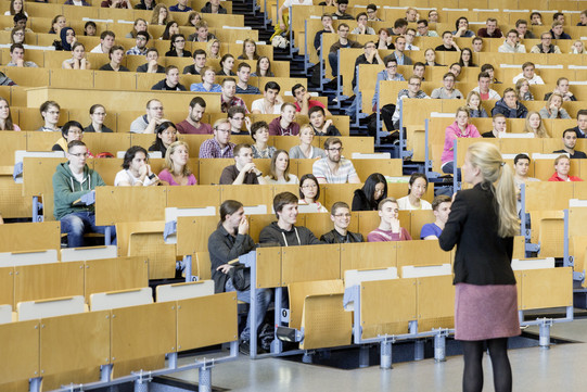 In einem Hörsaal sitzen viele Student*innen und vor ihnen steht die Dozentin