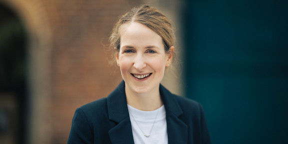 Ein Porträtfoto von Dr. Lena Tacke: Dr. Tacke hat blonde Haare und trägt einen schwarzen Blazer über einem weißen T-Shirt.