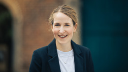 Dezember Ein Porträtfoto von Dr. Lena Tacke: Dr. Tacke hat blonde Haare und trägt einen schwarzen Blazer über einem weißen T-Shirt.