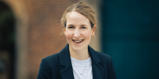 Ein Porträtfoto von Dr. Lena Tacke: Dr. Tacke hat blonde Haare und trägt einen schwarzen Blazer über einem weißen T-Shirt.