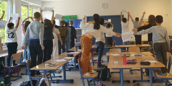 Das Bild zeigt auf den Stühlen stehende Schüler*innen, die einen DIN-A4 Blatt mit etwas Geschriebenem hochhalten. Die Schüler*innen wurden von der hinteren Wand des Raumes aus fotografiert.