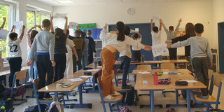 Das Bild zeigt auf den Stühlen stehende Schüler*innen, die einen DIN-A4 Blatt mit etwas Geschriebenem hochhalten. Die Schüler*innen wurden von der hinteren Wand des Raumes aus fotografiert.