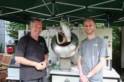 Foto: Zwei Männer stehen an einem Stand auf dem Sommerfest der TU Dortmund und lächeln in die Kamera. Hinter ihnen steht ein großer Apparat für einen Versuchsaufbau.