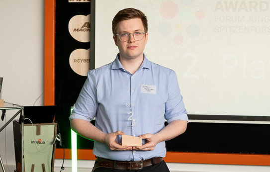 Foto: Ein Mann mit hellblauem Hemd und Brille steht vor einer Leinwand und hält eine Trophäe mit der Zahl 2 in der Hand.