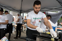 Eine Mann steht unter einem Pavillon und bedient ein Küchengerät. Im Hintergrund sind weitere Personen erkennbar. 