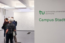 Das Bild zeigt auf der linken Seite die Weite des Ausstellungsraumes mit Besucher*innen und rechts im Bild ist auf weißer Wand das TU-Logo und darunter die Wörter "Campus Stadt" zu sehen.