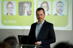 Das Bild zeigt einen Mann im Anzug, welcher hinter einem Rednerpult steht. Im Hintergrund ist eine Leinwand mit zwei grünen Kacheln und darin enthalten jeweils zwei Porträtfotos von Personen zu sehen.